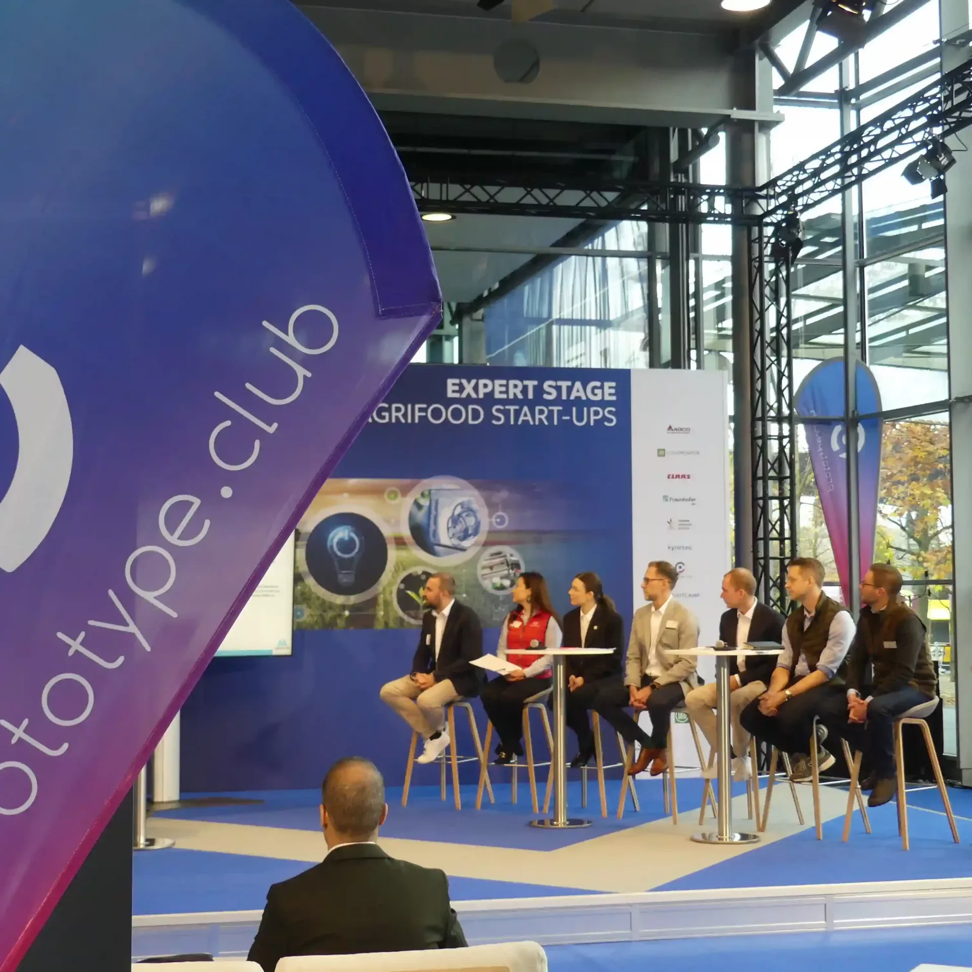 Bühne des Prototype.clubs mit der DLG. 7 Menschen auf der Bühnen hören sich gespannt den Pitch Vortrag eines Challenge-Teilnehmers an.