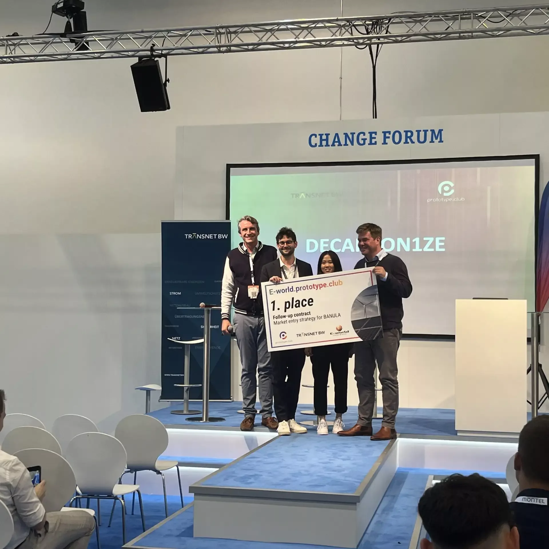 Preisvergabe auf der Bühne "Change Forum" . Die Gewinner halten einen Check mit dem ersten Platz in die Kamera. Auf der Bühne stehen Vertreter von prototype.club, TransnetBW und die Gewinner Decarbon1ze.
