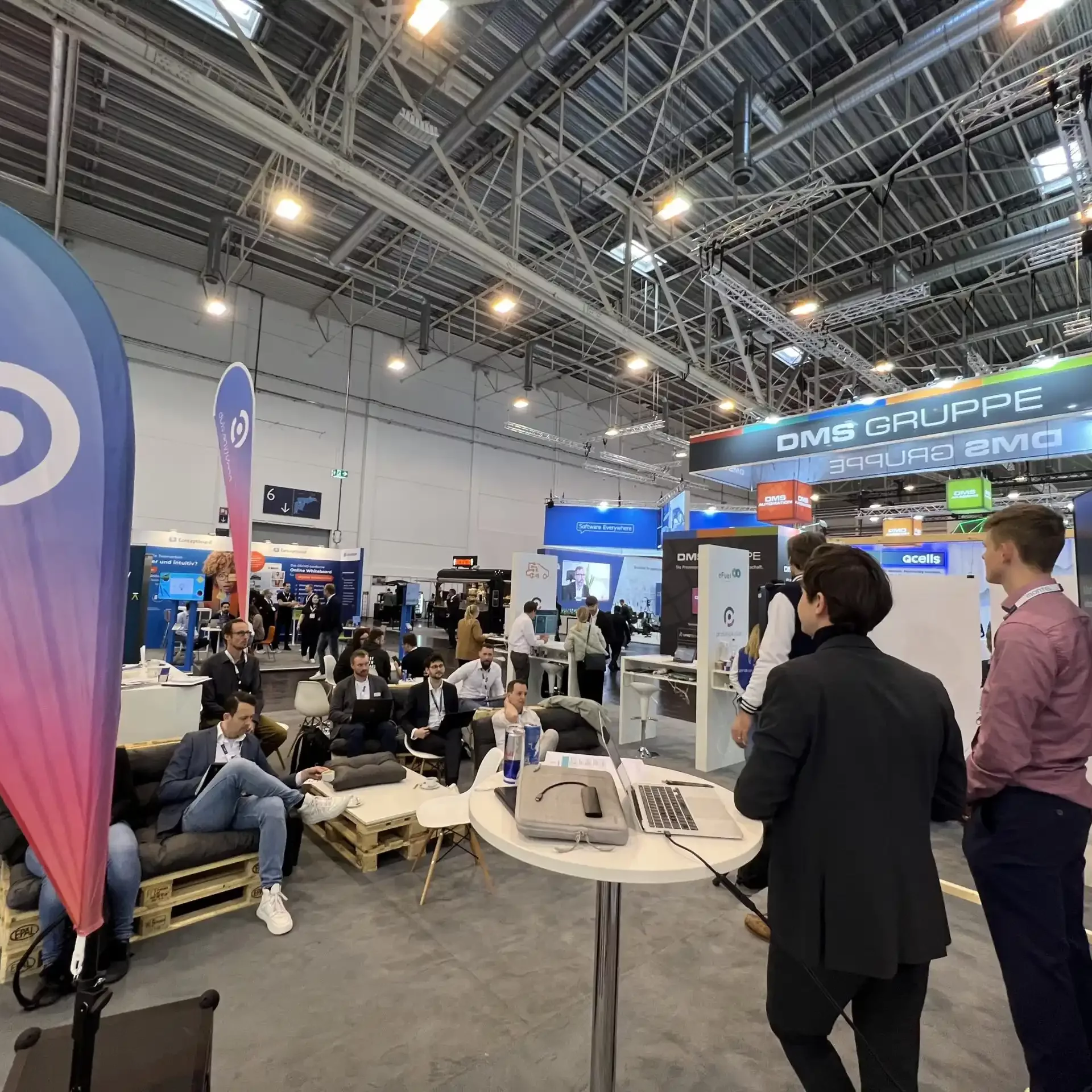 Pitch eines Startups an dem Messestand von prototype.club. Es wird in das Publikum fotografiert. Man sieht die Vortragenden von hinten links. Links im Vordergrund weht eine blau-lila Beachflag des prototype.club.